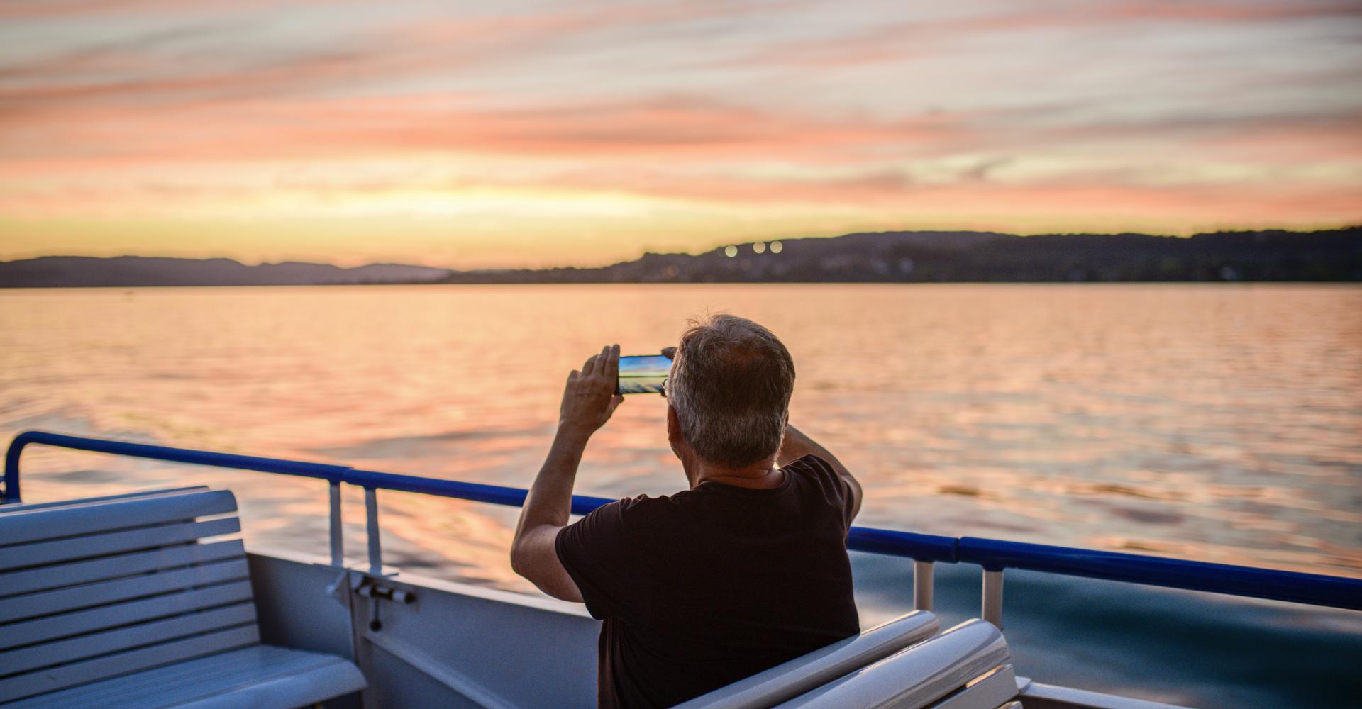 Mann sitz im Schiff auf dem Zürichsee bei Abendstimmung und macht ein Bild