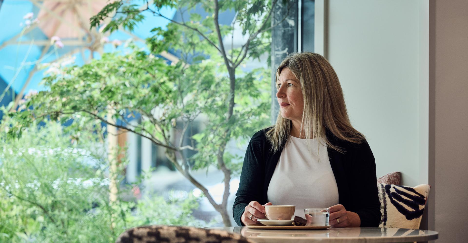 Frau sitzt an einem Tisch beim Fenster und trinkt Kaffee