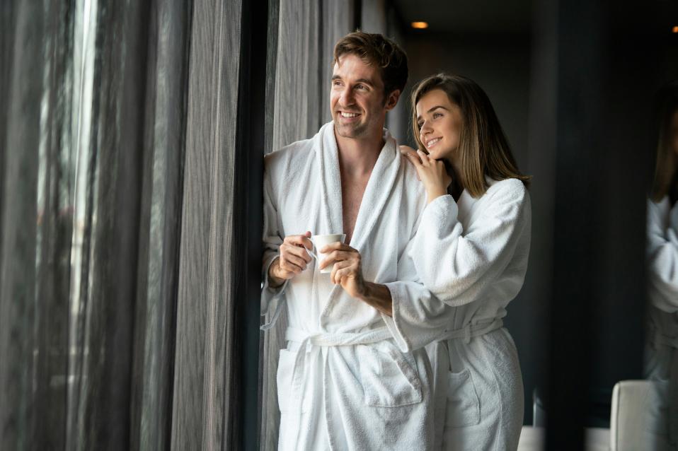 Paar im Bademantel steht im Hotelzimmer am Fenster