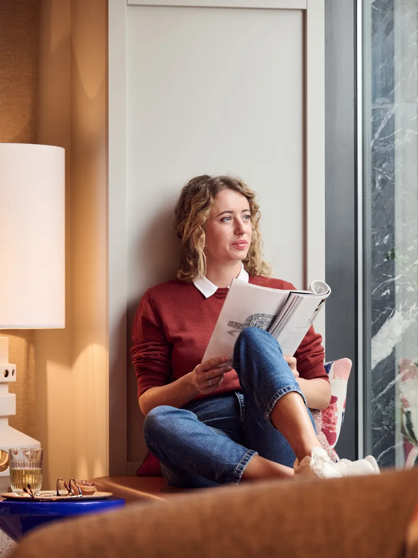 Frau liest ein Magazin auf einem Fenstersims und blickt durch ein Fenster