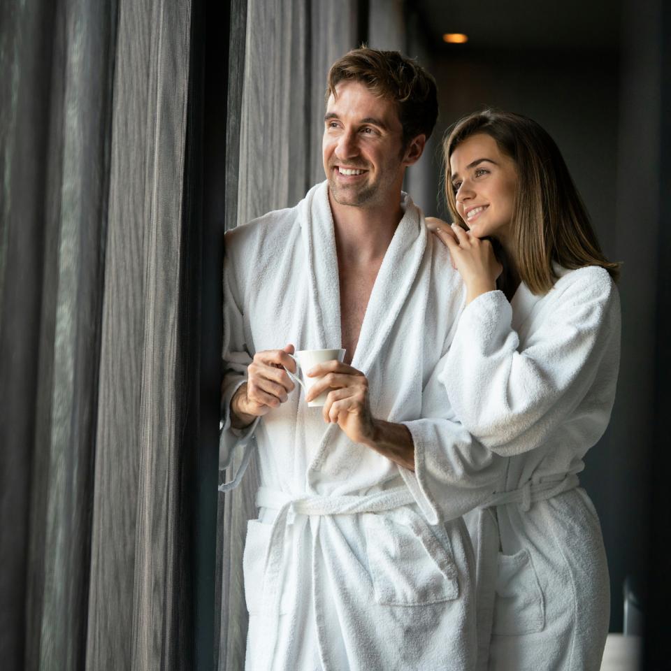 Paar im Bademantel steht im Hotelzimmer am Fenster