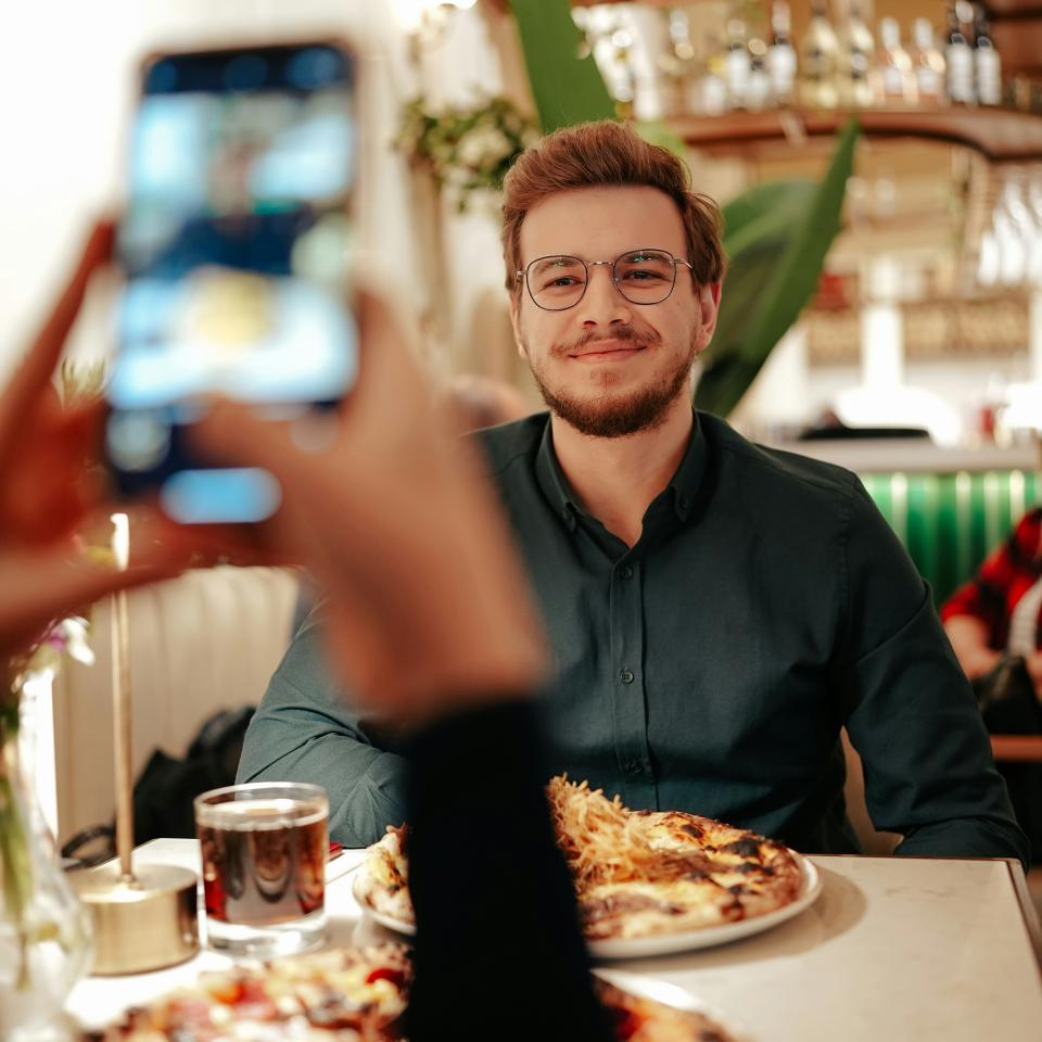 Mann sitzt am Tisch vor Essen und eine Frau mach ein Bild von ihm