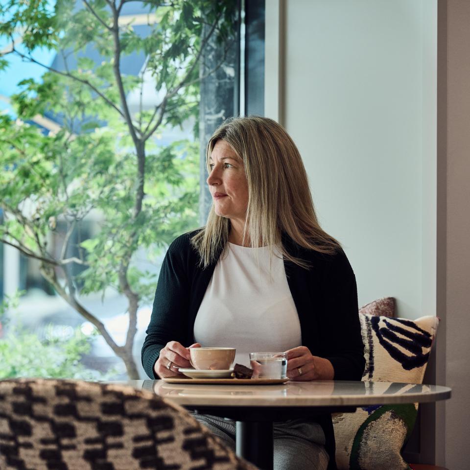 Frau sitzt an einem Tisch beim Fenster und trinkt Kaffee